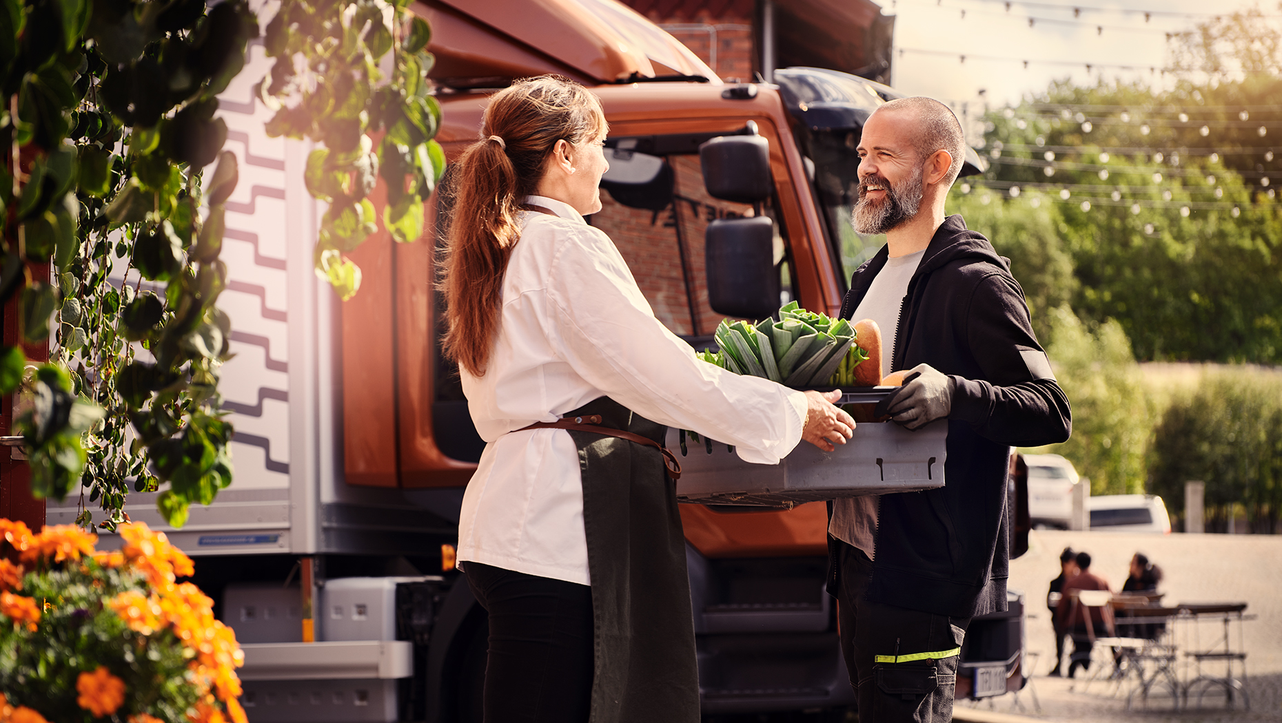 Smiling man handing over a box of vegetables to a woman wearing an apron with a Volvo Truck in the background