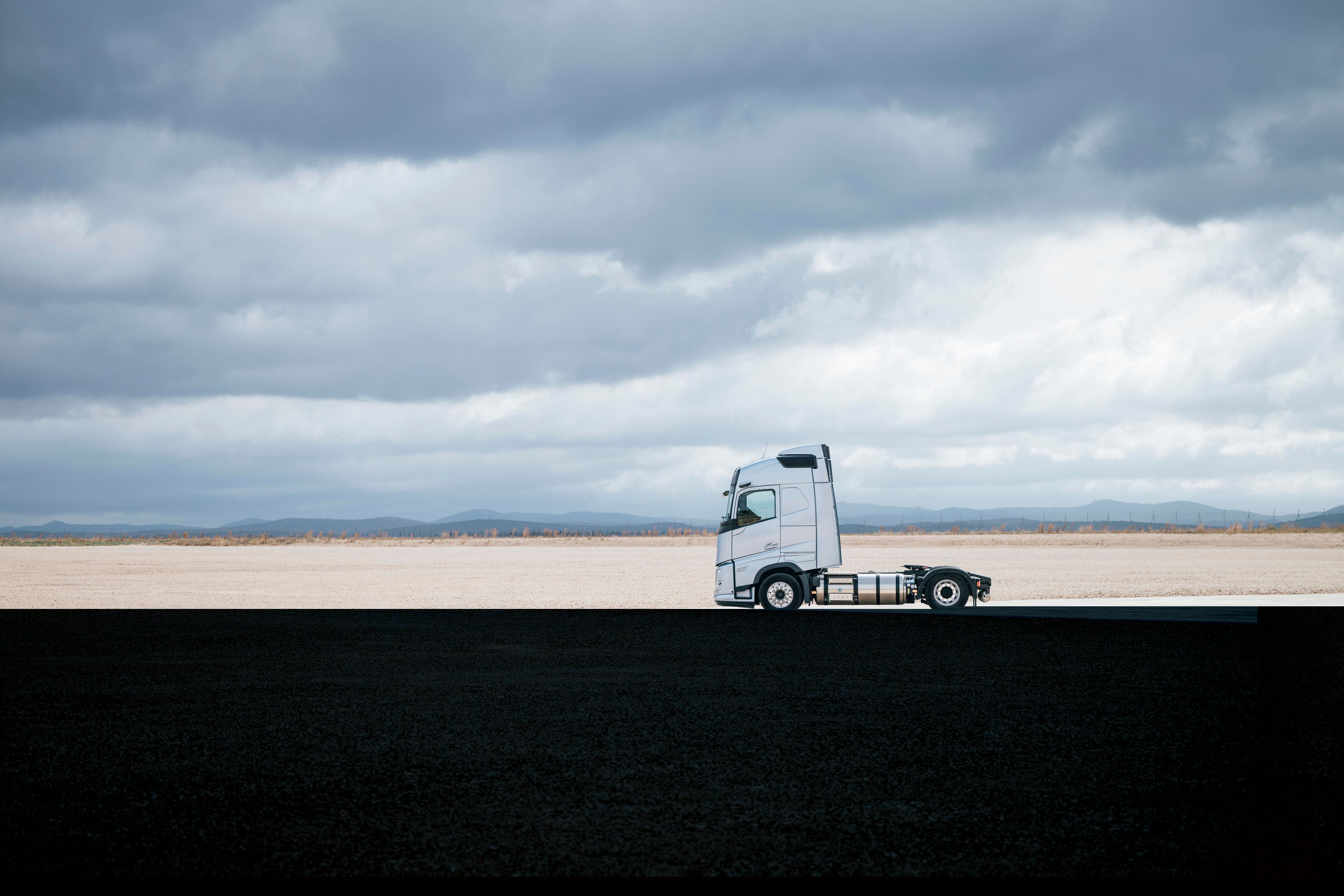 Zijaanzicht van een Volvo-vrachtwagen die geparkeerd staat in een weids, open landschap onder een bewolkte hemel, met een uitgestrekte omgeving en een minimalistische achtergrond.