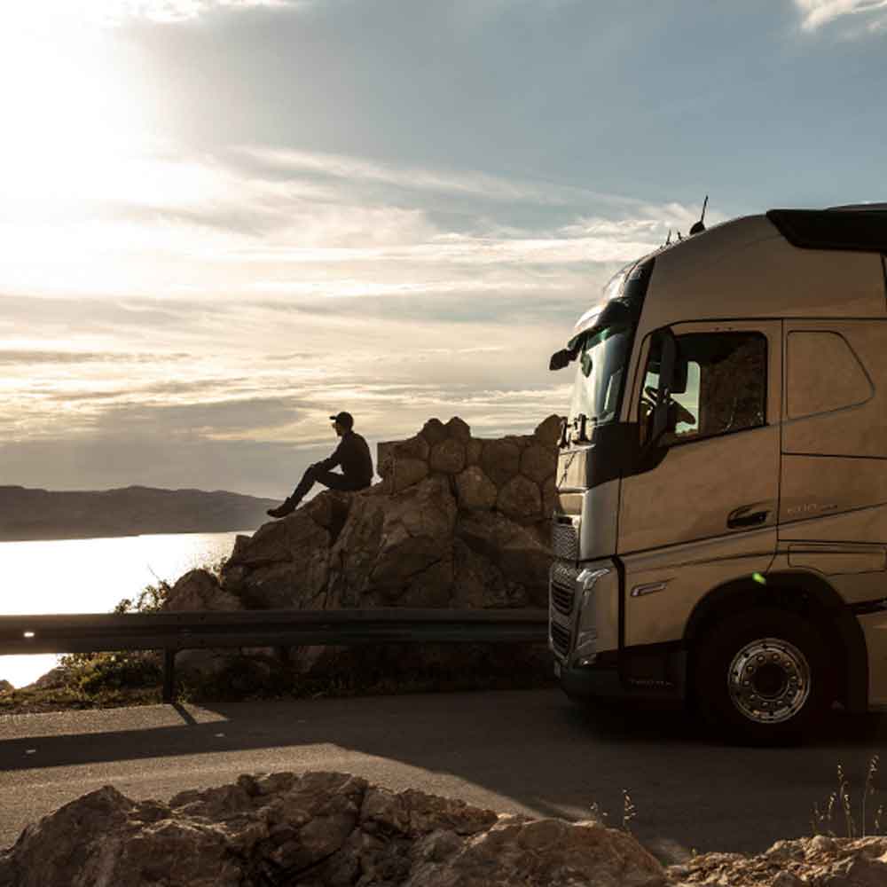 Een chauffeur zit op een rots met uitzicht op zee bij zonsondergang, met zijn Volvo FH-truck naast hem geparkeerd